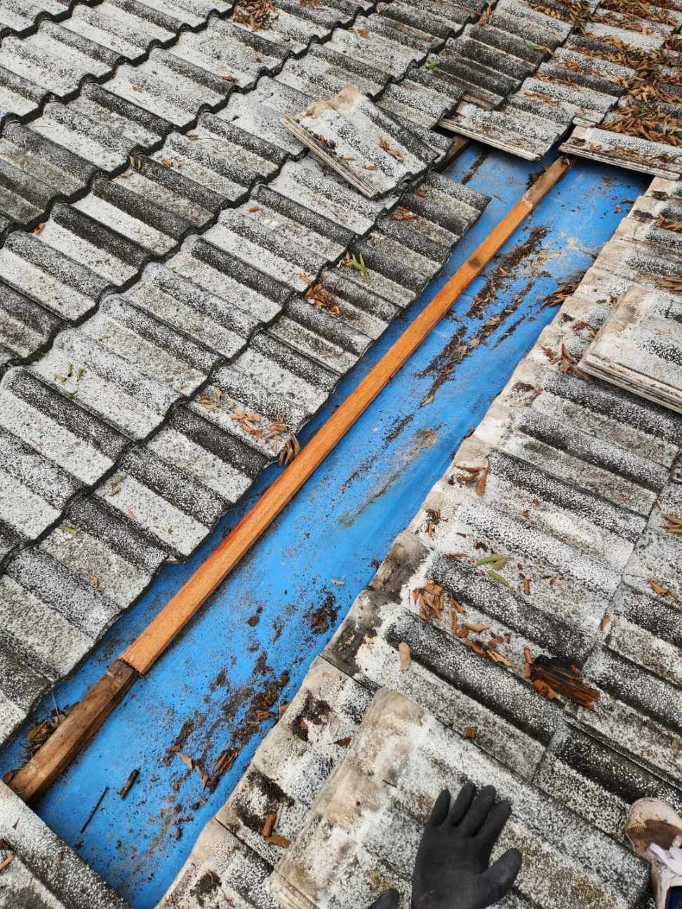 A patch of grey roof tiles removed to reveal a wooden beam over blue underlayment, viewed from above by a gloved hand.