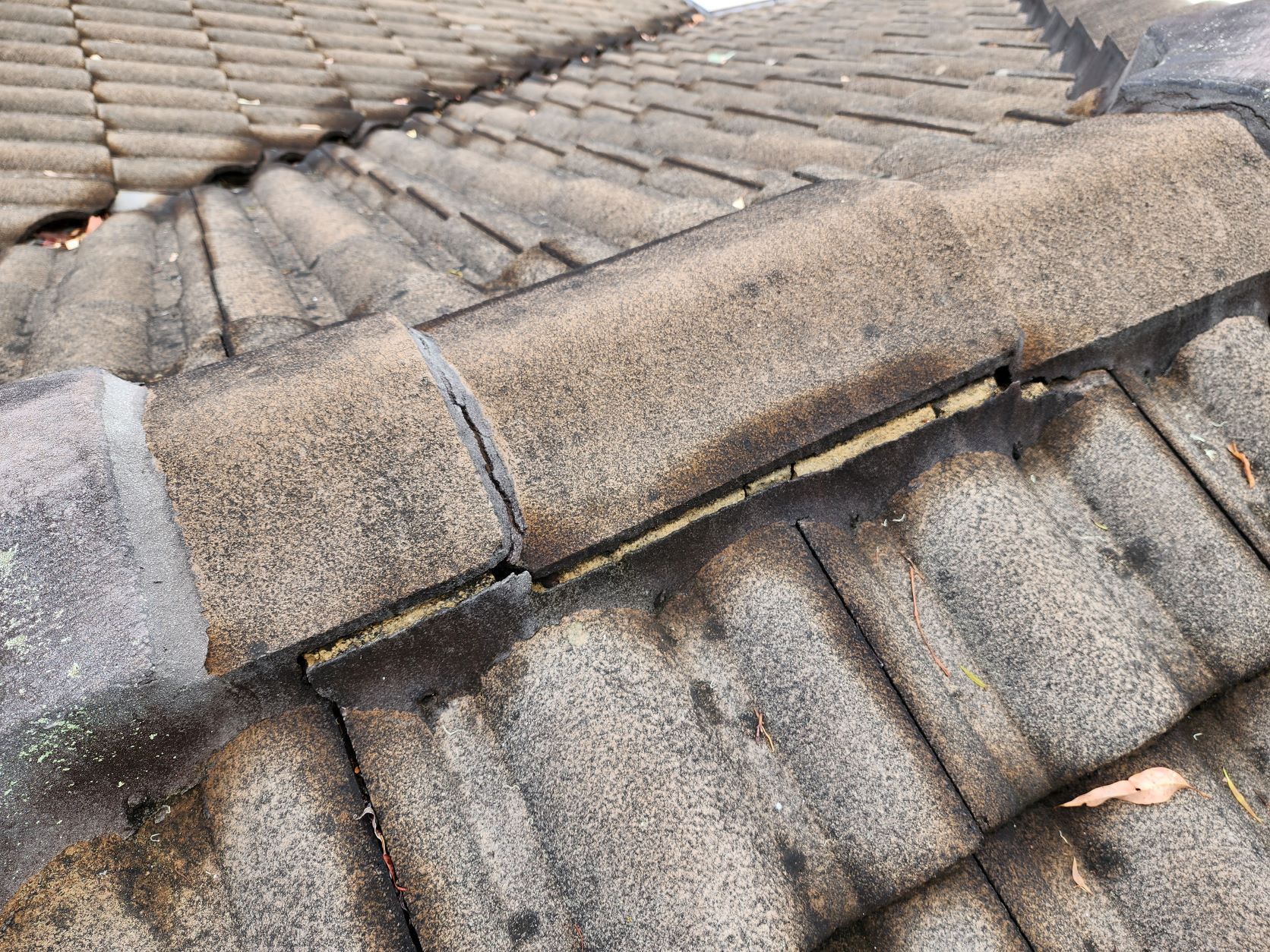 A close-up of a weathered, grey tiled roof showing gaps and cracking in the mortar between the ridge tiles.