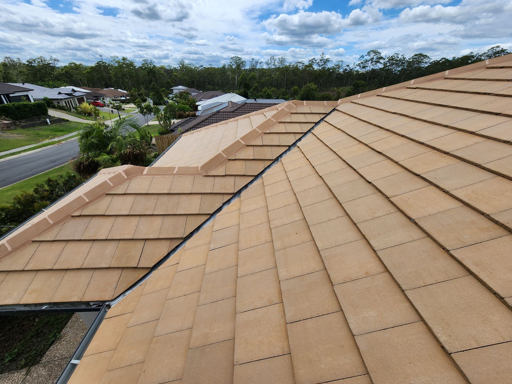 A high-angle view of a beige tiled roof featuring a hip and valley design, overlooking a suburban street and trees.