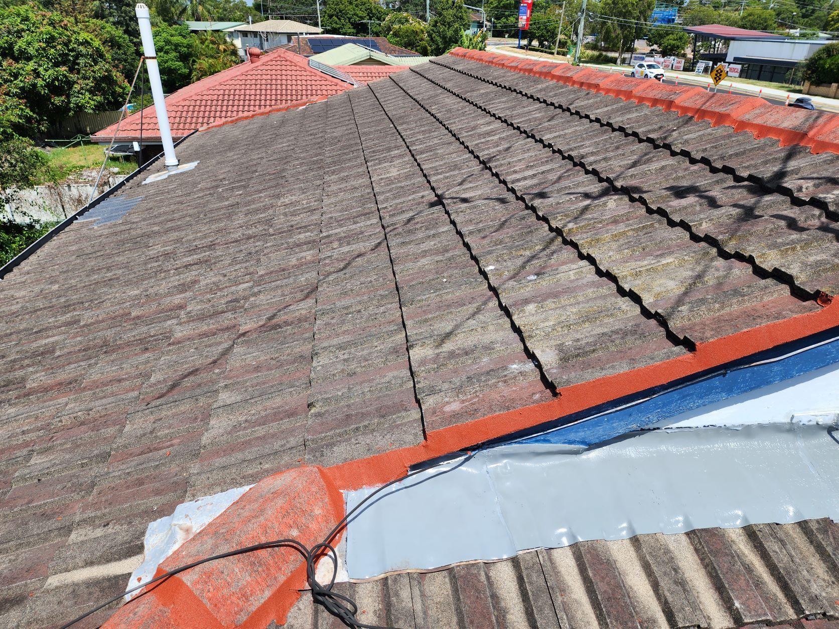 A high-angle view of a weathered, dark-tiled roof with a red ridge cap and a section of new silver flashing.