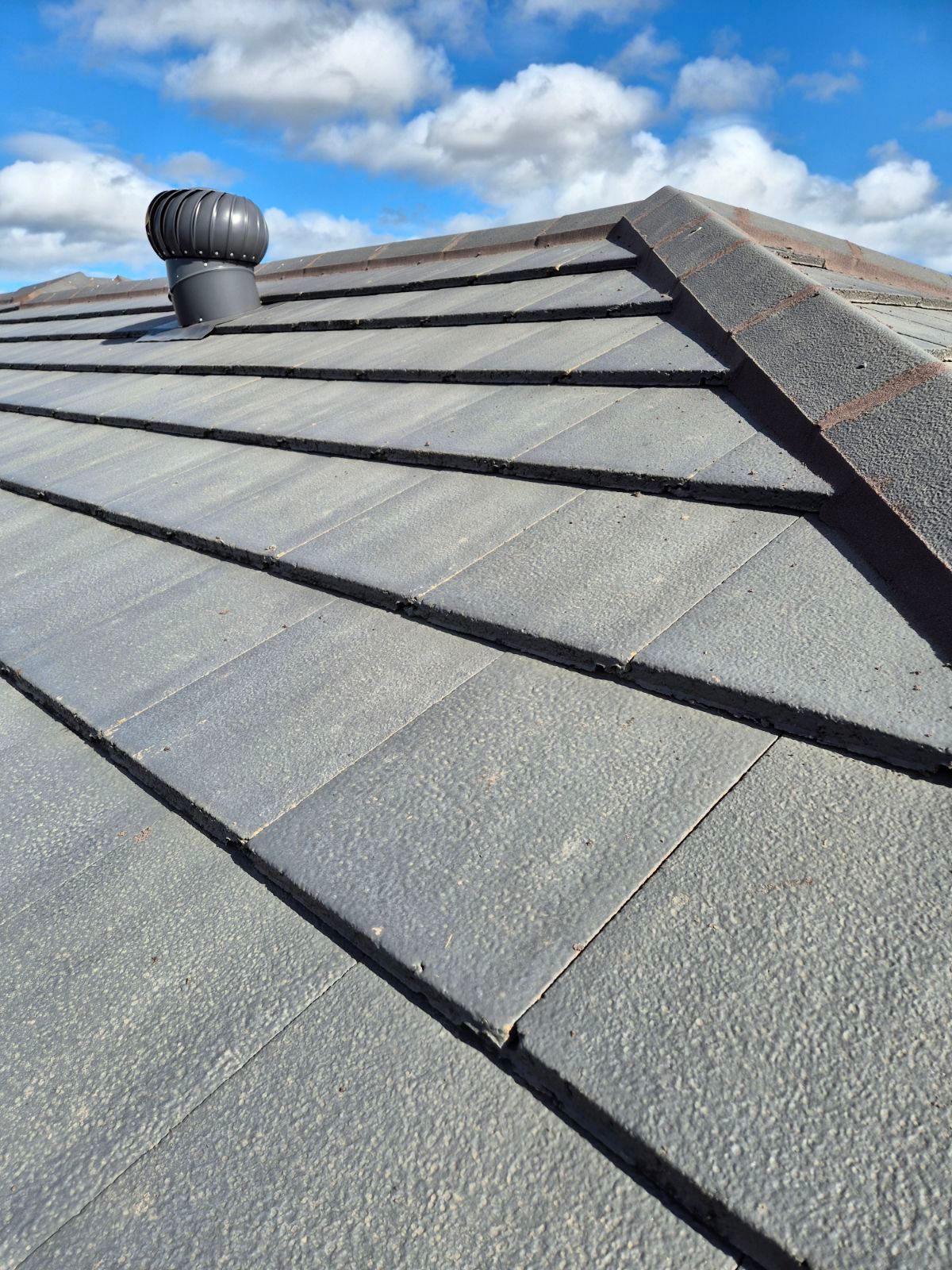 A gray tiled roof featuring a dark ridge cap and a roof ventilation fan under a bright blue sky with scattered clouds.