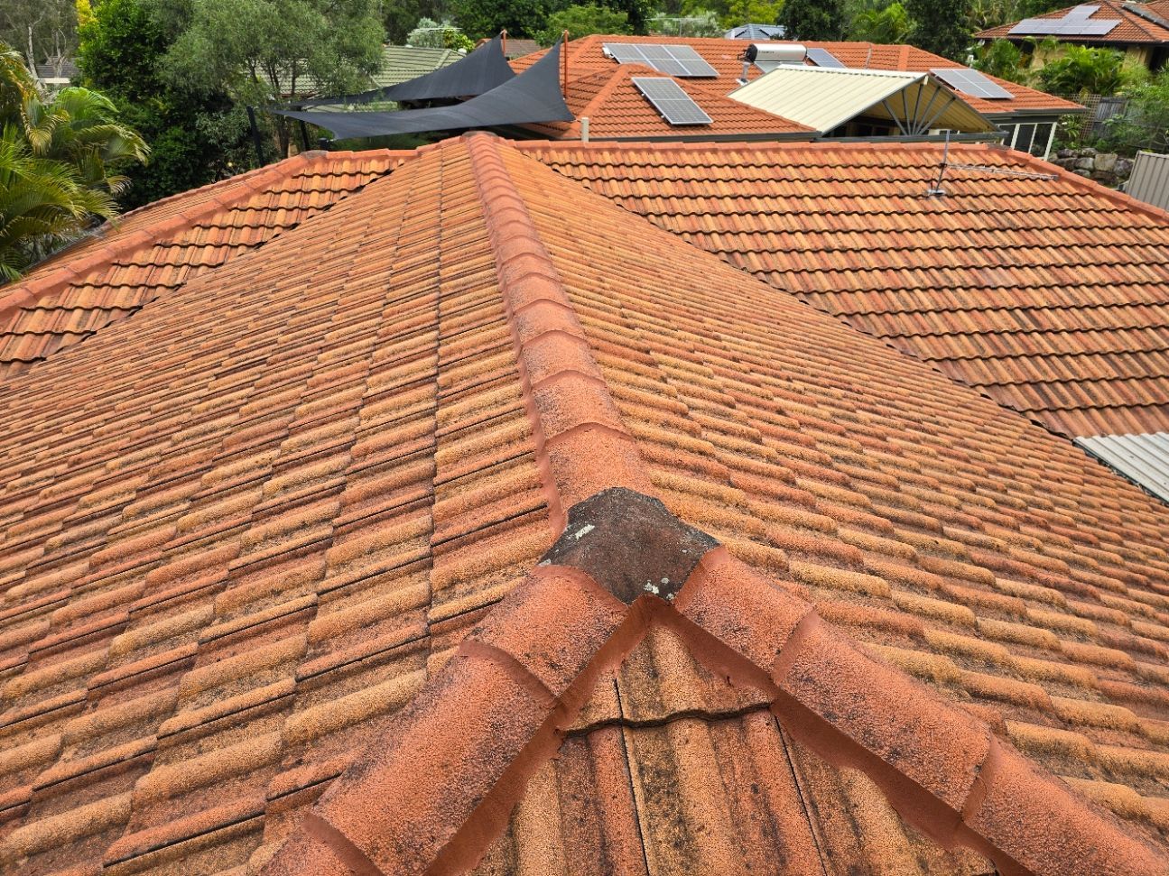An aerial view of a textured, terra cotta tiled residential roof with a prominent ridgeline and solar panels in back.