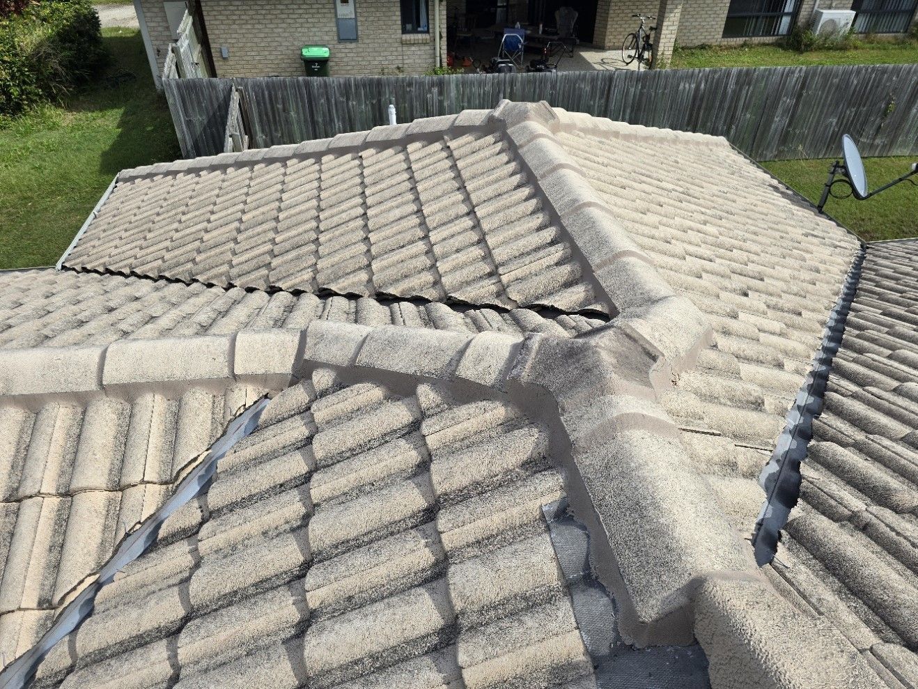 High-angle view of a tiled roof with ridges and valleys, showing light gray textured tiles and a satellite dish.