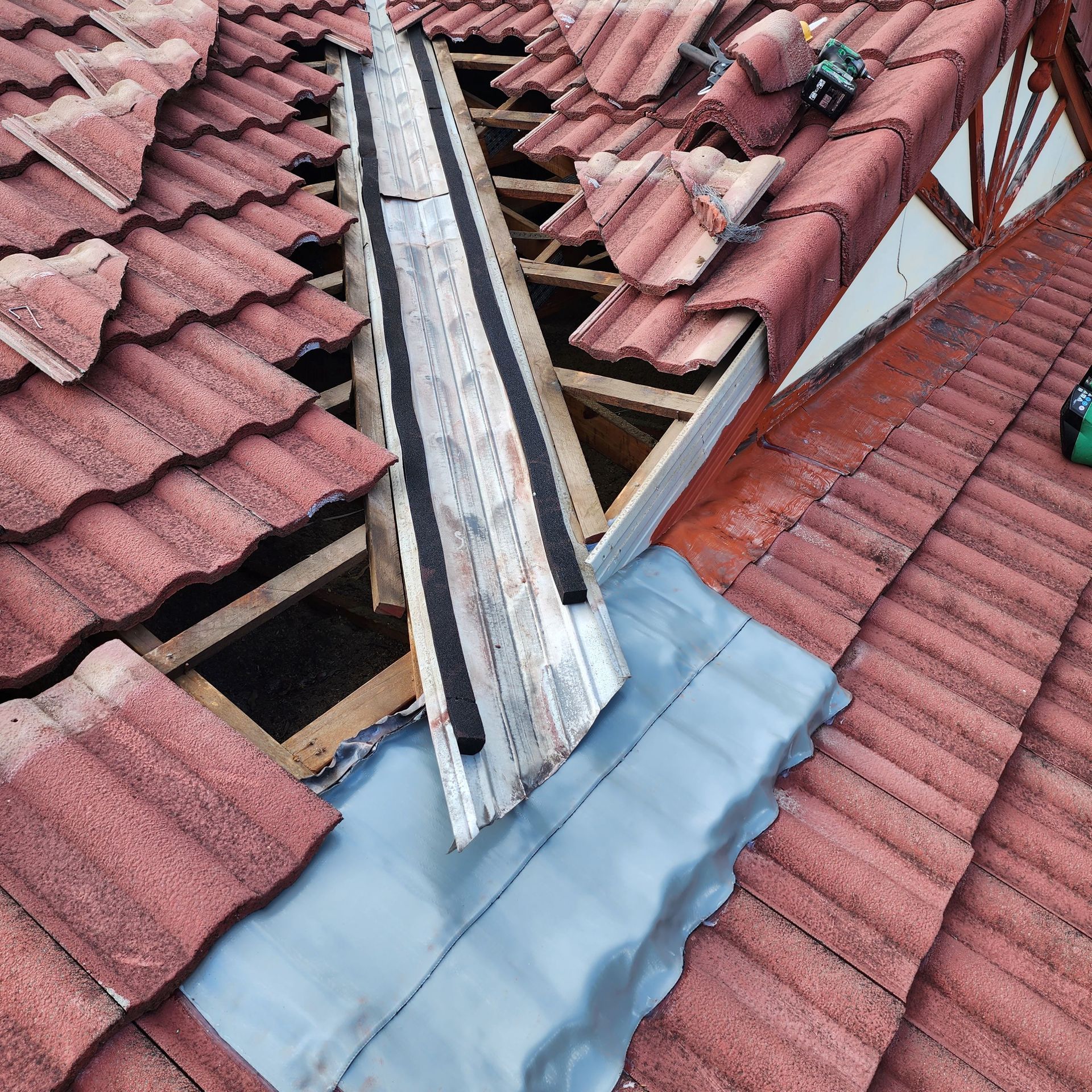 A roof valley being repaired, showing exposed wooden rafters, a metal flashing channel with black foam, and gray underlay.