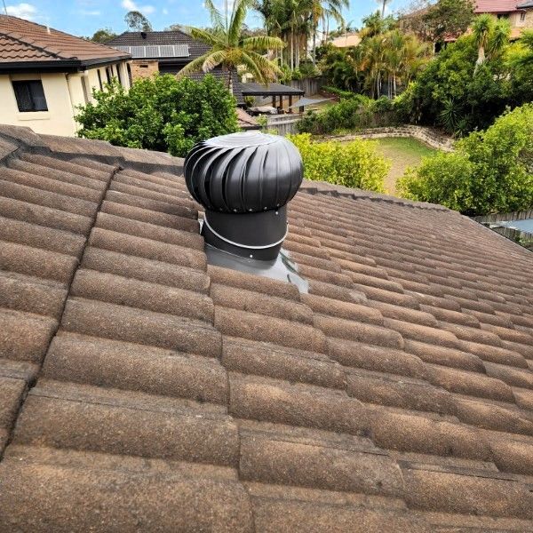 A black roof turbine vent mounted on a brown tiled roof, with residential houses and trees in the background.