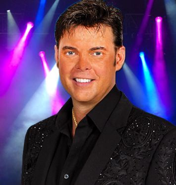 A man in a black suit is smiling in front of a stage with purple and blue lights.