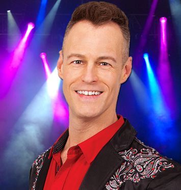 A man in a red shirt and black jacket is smiling on a stage.