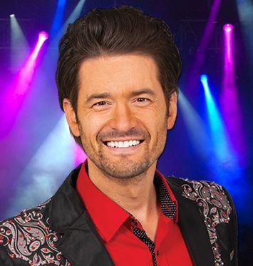A man in a red shirt and black jacket is smiling in front of a stage.