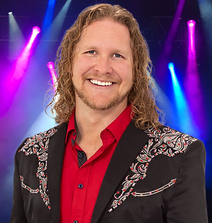A man with long hair and a beard is wearing a red shirt and a black jacket.