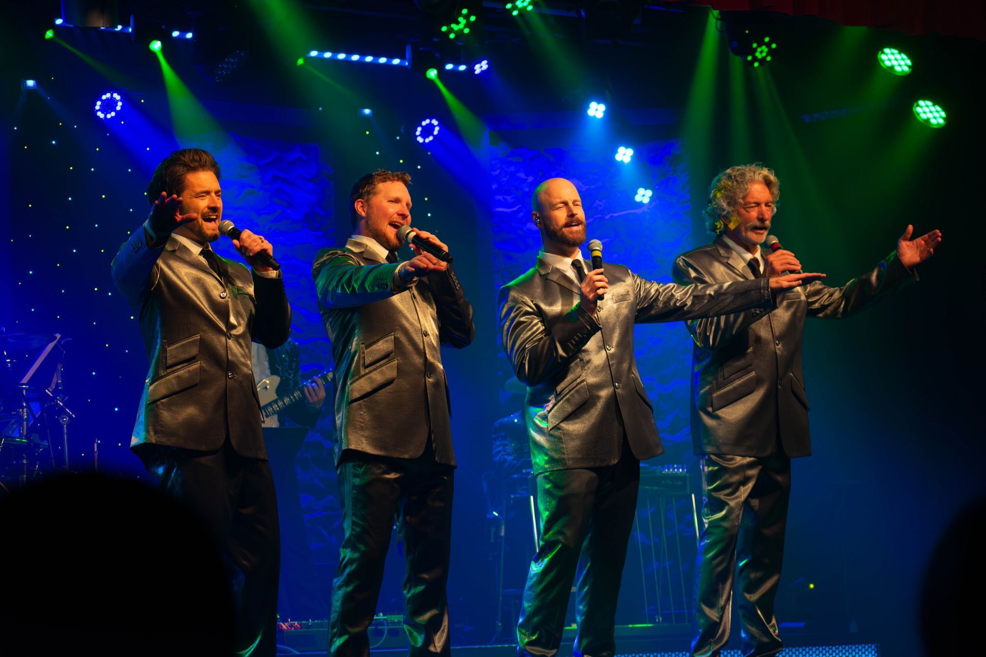 Four men in suits are singing into microphones on a stage.