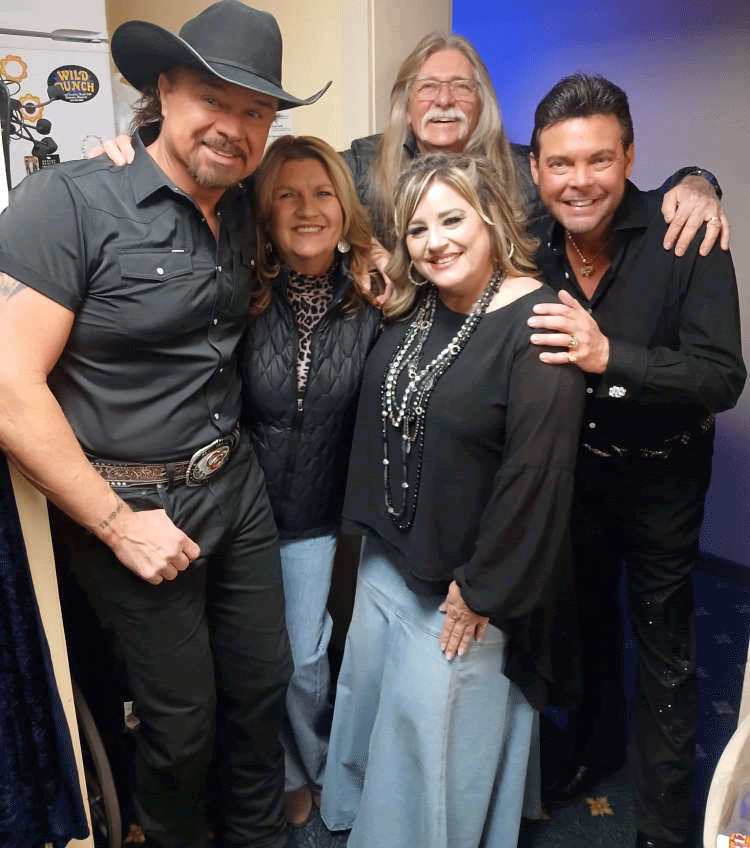Group of people smiling, posing for a photo. Men in black shirts, a cowboy hat, and two women in casual attire.