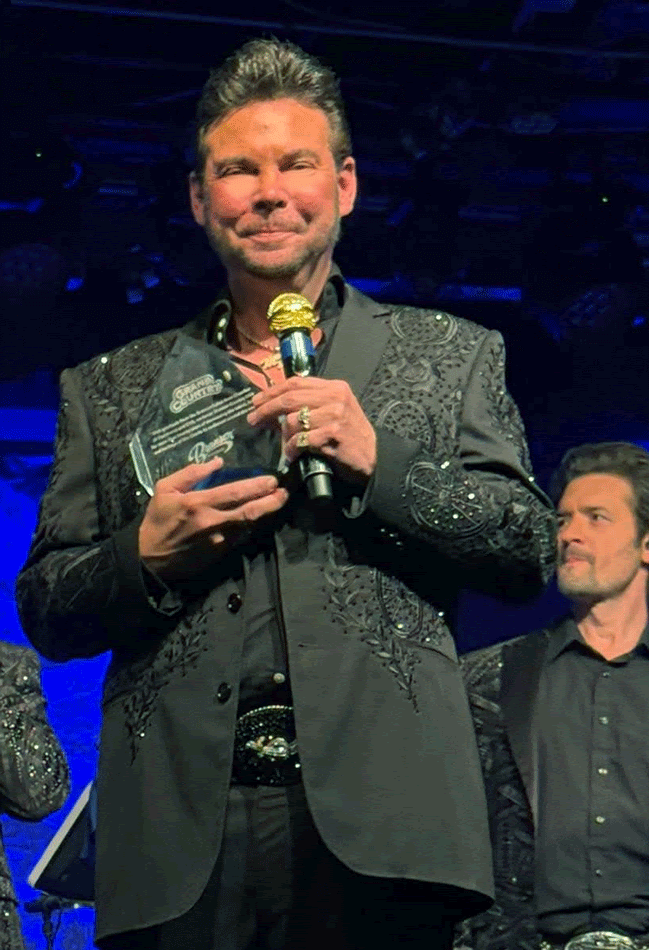 Man in black jacket holding an award and microphone on stage. Smiling, with another man in background.