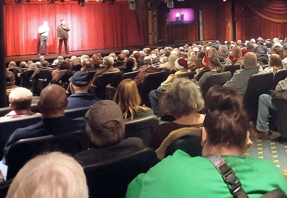 Audience seated, watching two men on stage with a red curtain backdrop. Dim lighting.