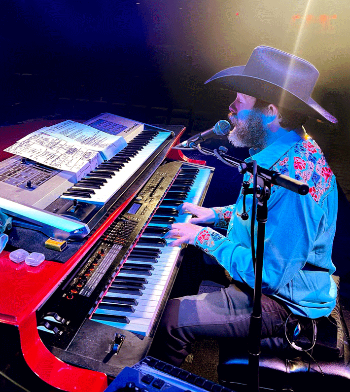 A group of costumed performers posing onstage with a snowman prop. Christmas theme, festive outfits, and smiling faces.