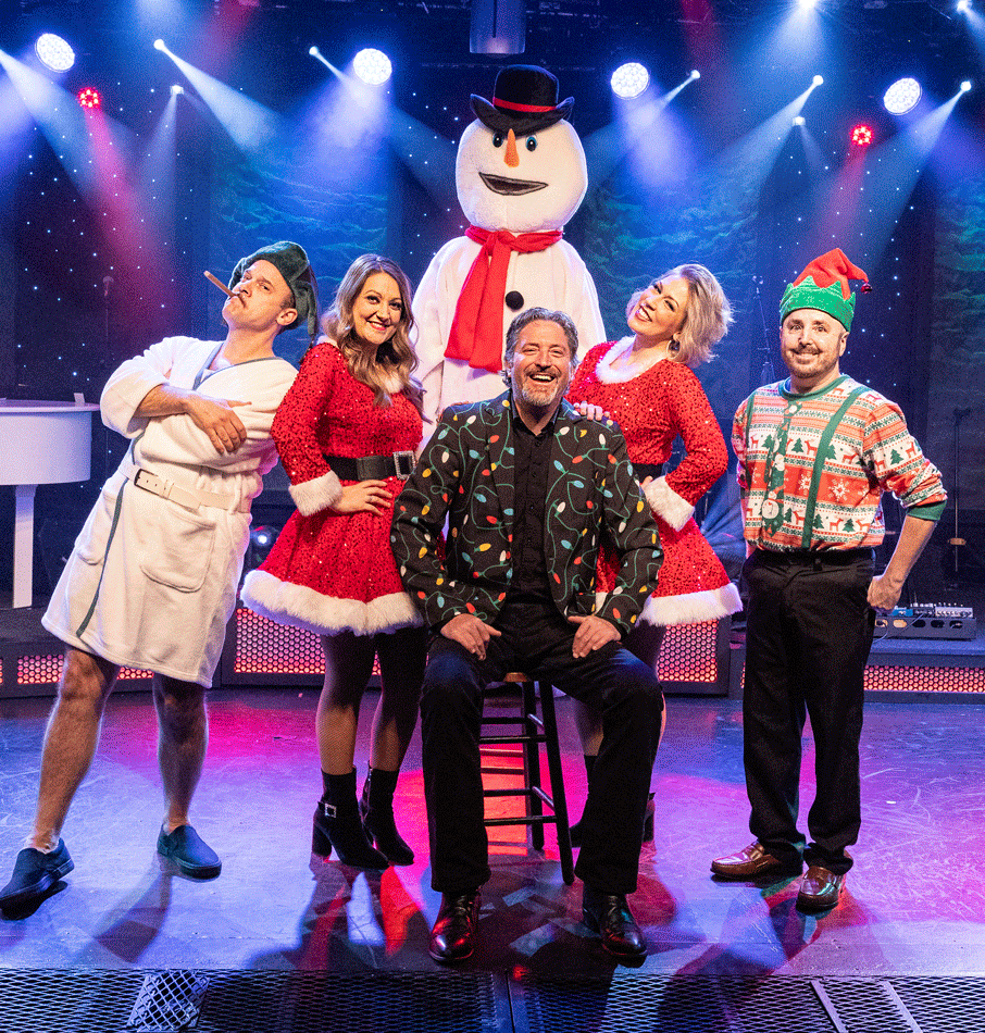 A group of costumed performers posing onstage with a snowman prop. Christmas theme, festive outfits, and smiling faces.