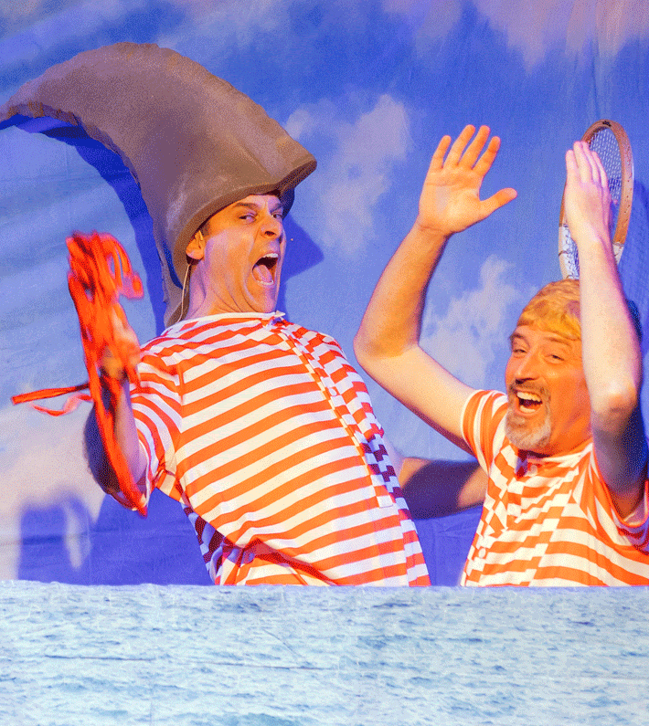 A group of costumed performers posing onstage with a snowman prop. Christmas theme, festive outfits, and smiling faces.
