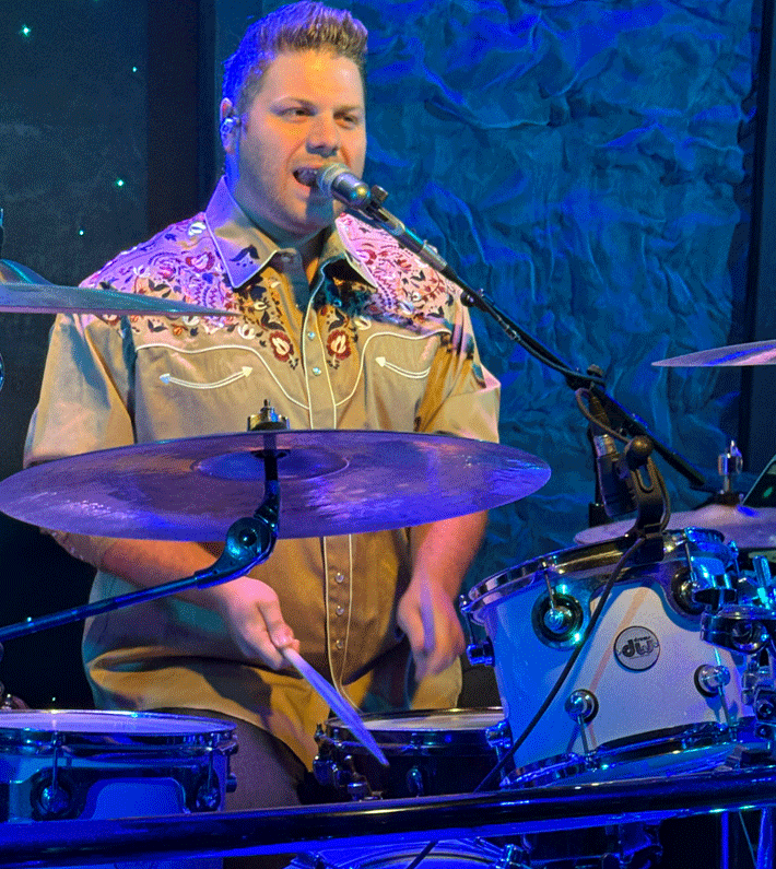 A group of costumed performers posing onstage with a snowman prop. Christmas theme, festive outfits, and smiling faces.