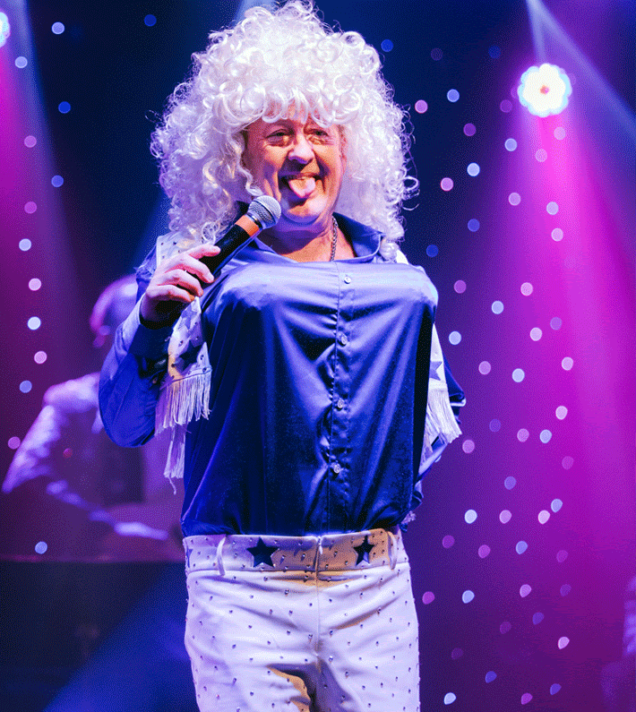 A group of costumed performers posing onstage with a snowman prop. Christmas theme, festive outfits, and smiling faces.