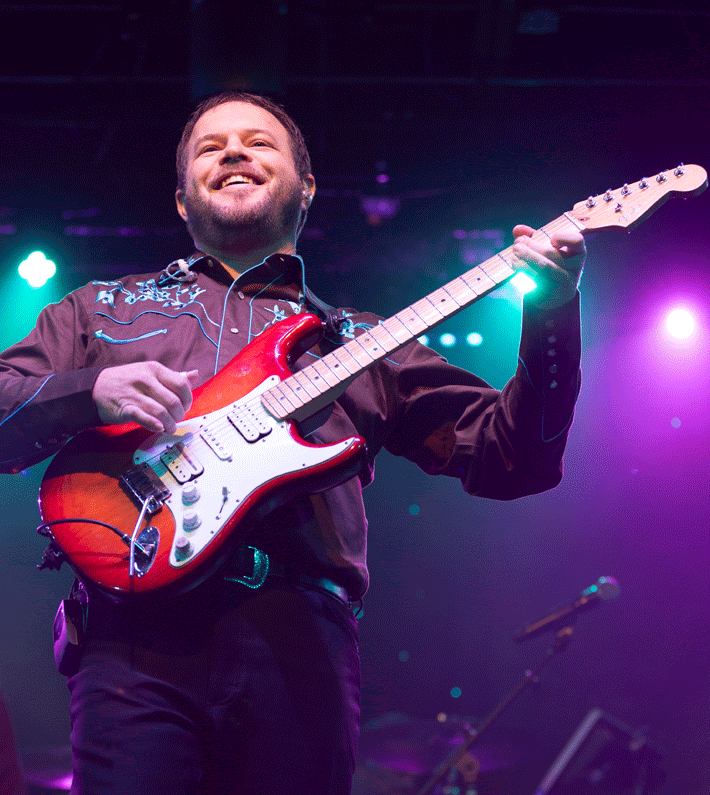 Smiling musician playing a red electric guitar on a purple-lit stage
