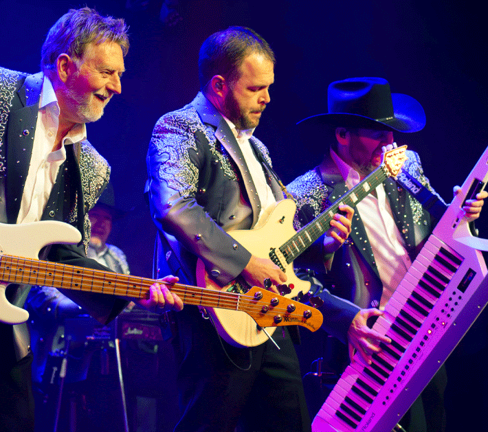 Smiling musician playing a red electric guitar on a purple-lit stage