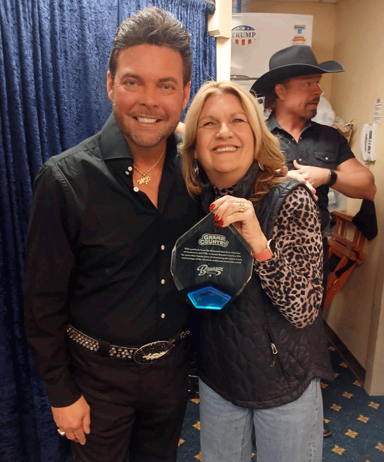 Man and woman smiling, holding an award backstage. Man wears black, woman wears a vest and jeans. A man in a cowboy hat is in the background.