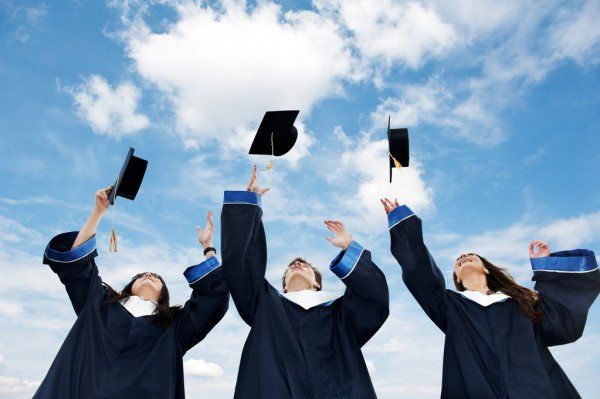 Graduates in caps and gowns toss their mortarboards into a blue sky outdoors