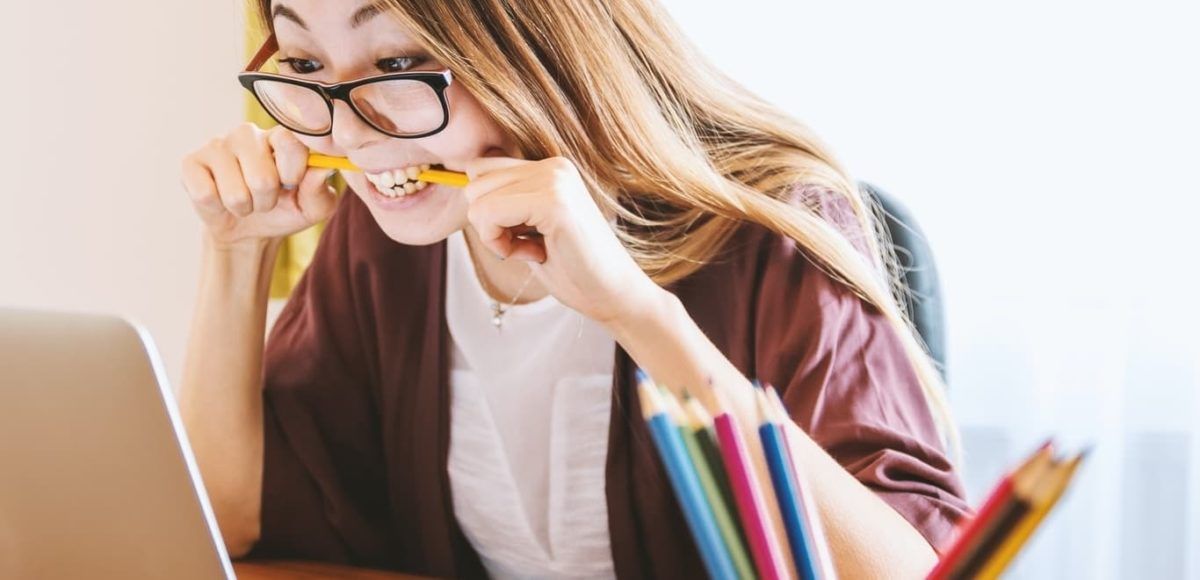 Woman wearing glasses, biting a pencil, looking at a laptop with a stressed expression. Colored pencils are on the table.