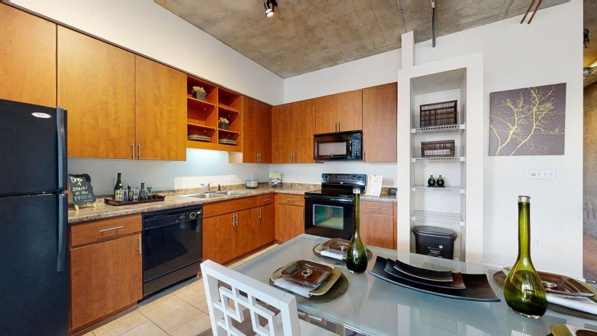 Modern industrial kitchen and living space here at Skyline Lofts Apartments with exposed ductwork and concrete ceiling. Features wood cabinets, black appliances, a central island with bar stools, and large windows offering natural light. Cozy seating area with patterned chairs and minimal decor.