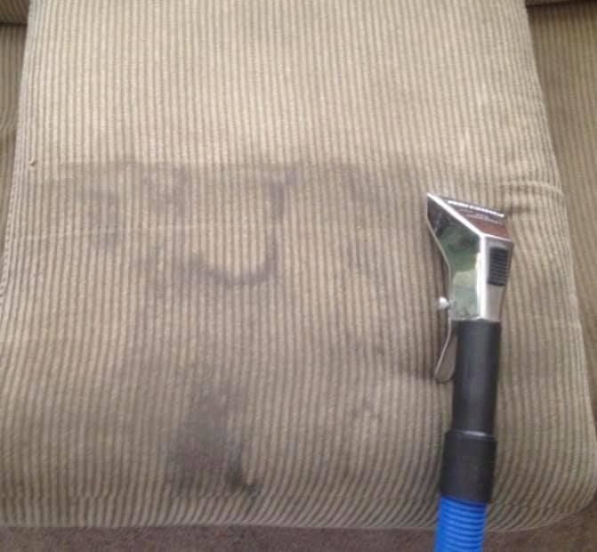 A fabric couch cushion with dark stains being cleaned by a handheld vacuum attachment.
