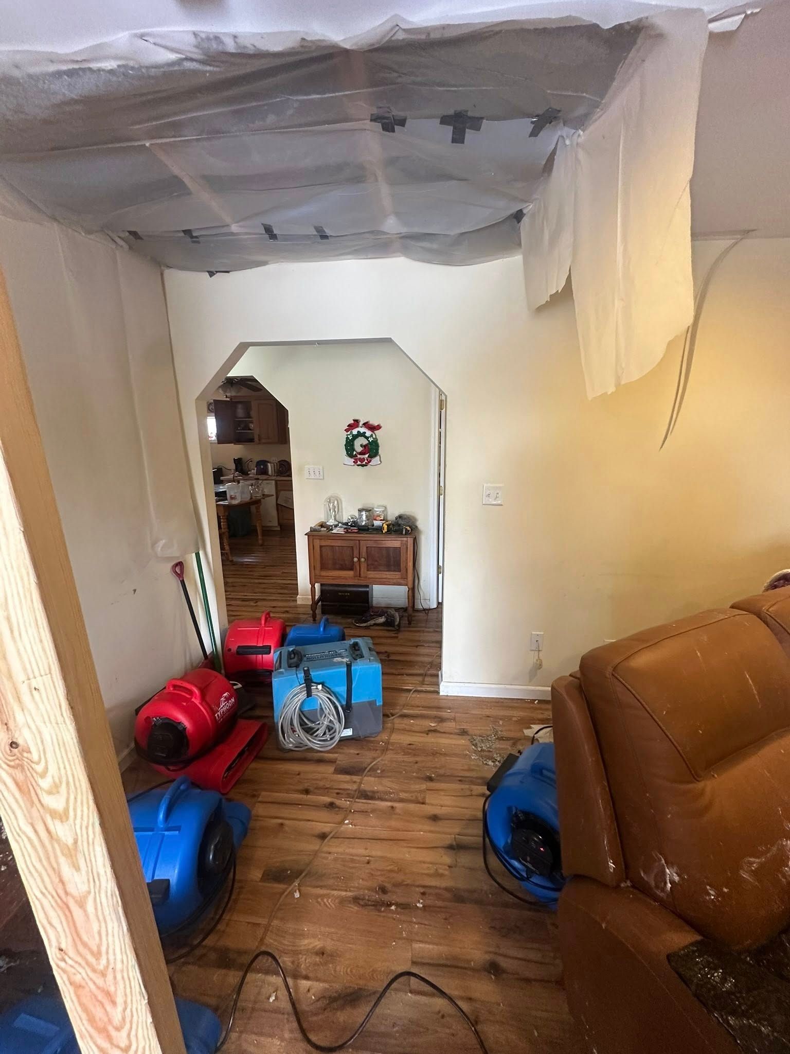 Room with water damage, plastic sheeting on ceiling, drying equipment on floor, doorway leads to another room.