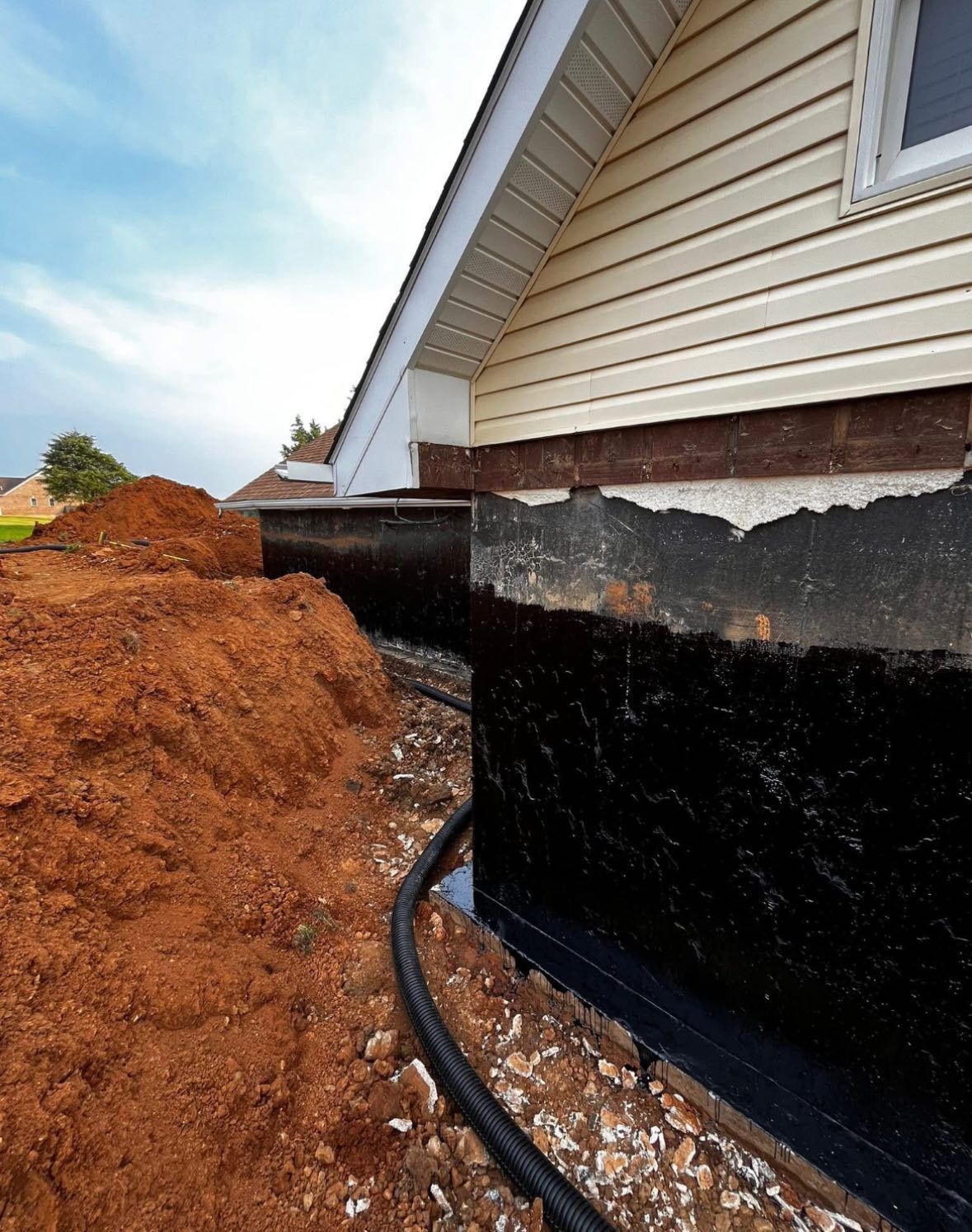 House foundation being waterproofed with black sealant after excavation. Reddish dirt pile, gravel