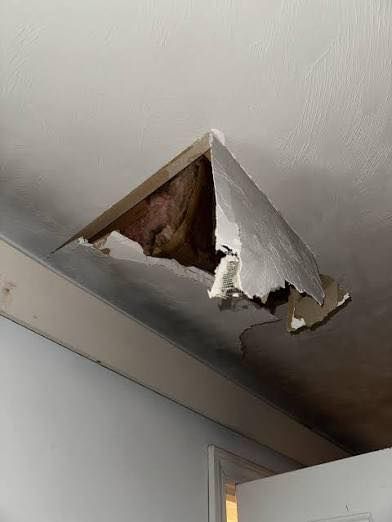 Damaged ceiling with a large hole revealing wood and insulation. White ceiling with peeling drywall.
