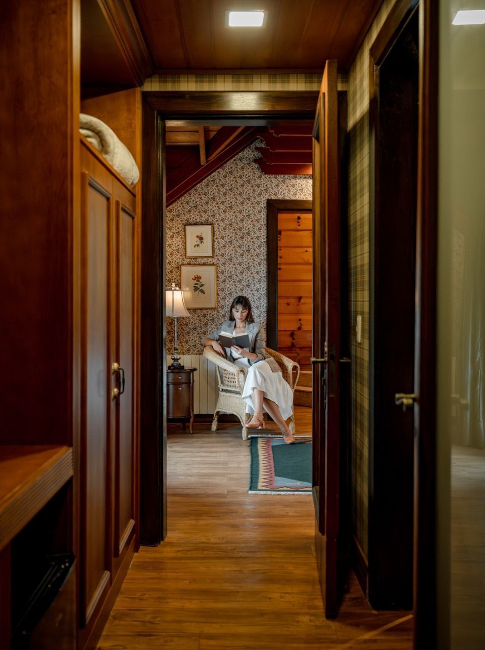Pessoa sentada em um quarto aconchegante, vista através de um corredor de madeira escura com iluminação quente.