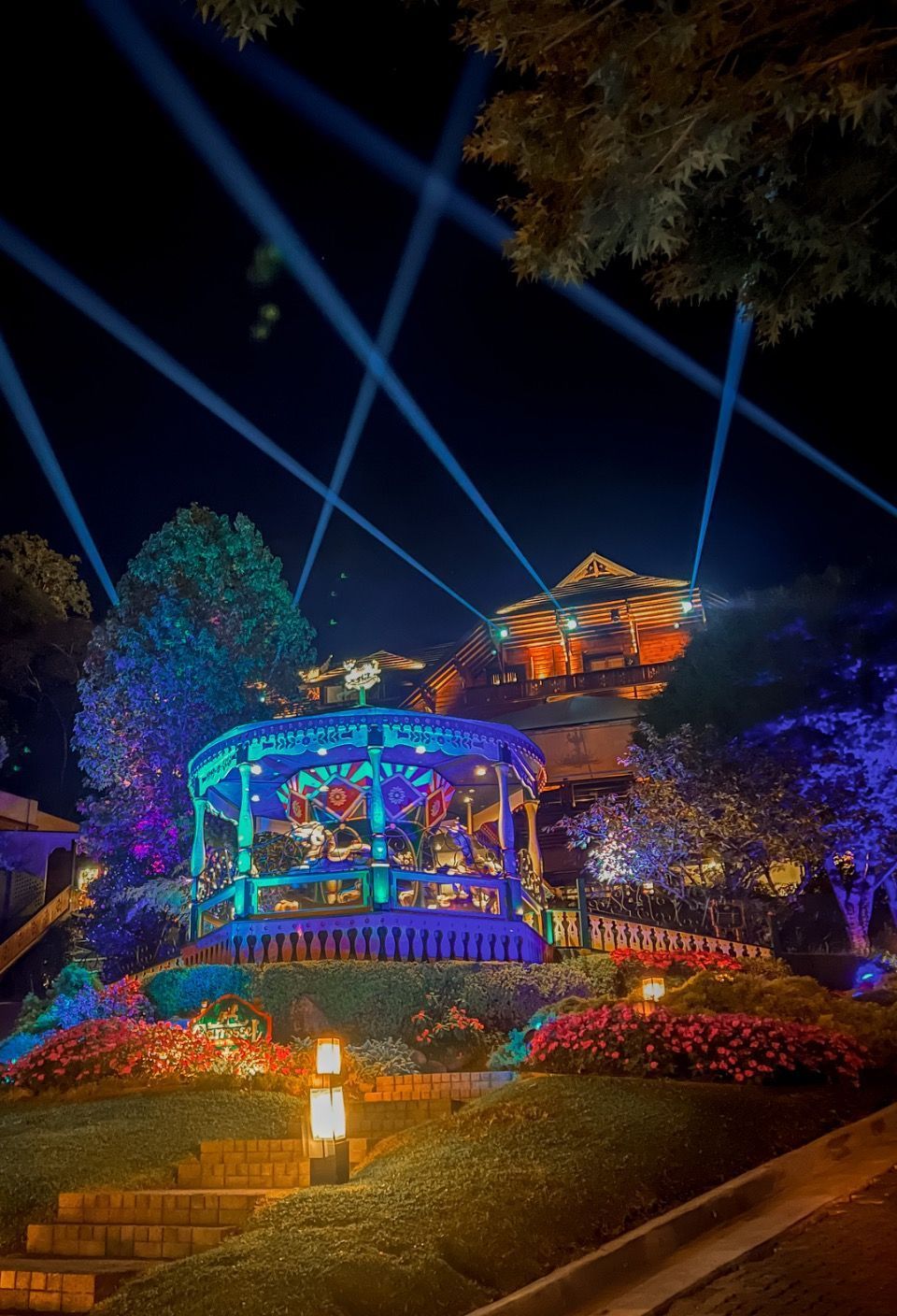 Pavilhão iluminado e colorido à noite com feixes de laser azuis, flores brilhantes e um jardim na encosta.