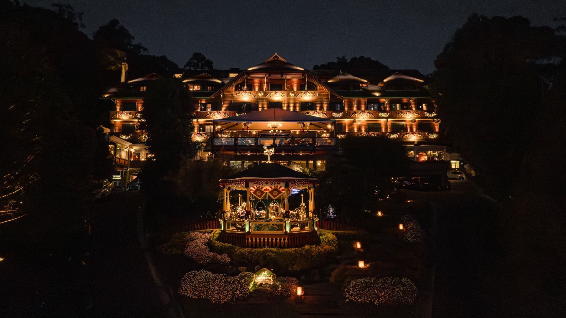 Vista noturna de uma casa de vários andares com iluminação aconchegante, cercada por árvores escuras e luzes de jardim.