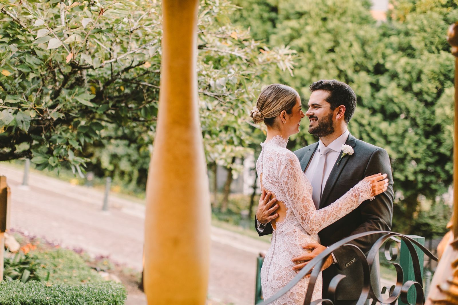 Casal em trajes formais se abraçando em um jardim, com árvores verdes e um caminho ao fundo.