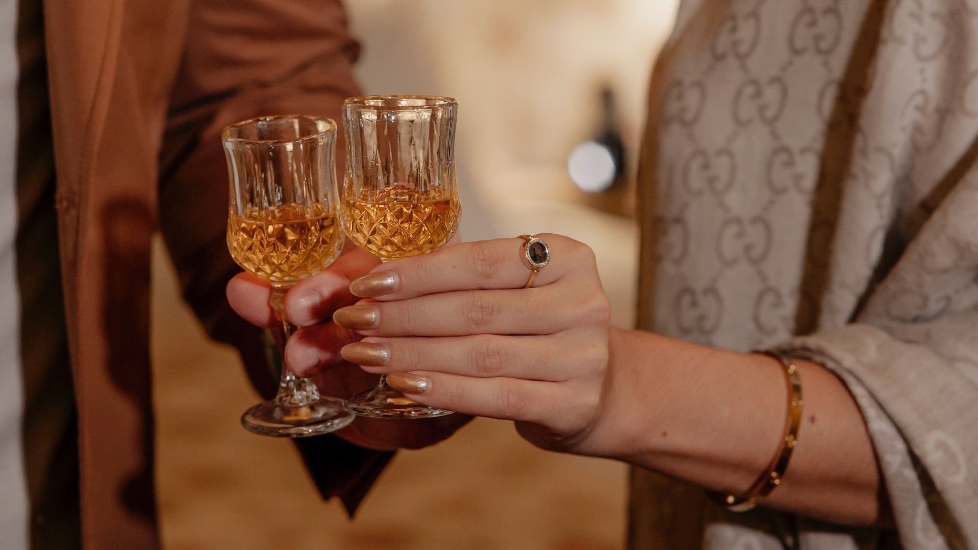 Duas mãos tilintam taças de cristal com bebida âmbar em uma reunião formal em ambiente fechado.