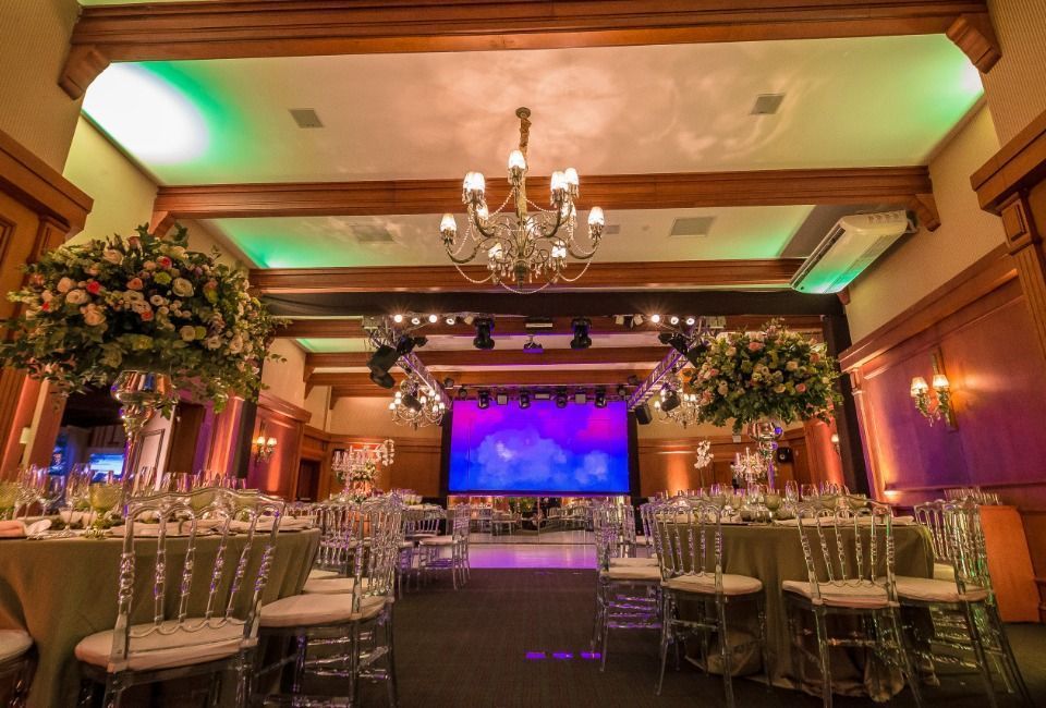 Elegante salão de banquetes com lustres, arranjos florais e um palco iluminado ao fundo.