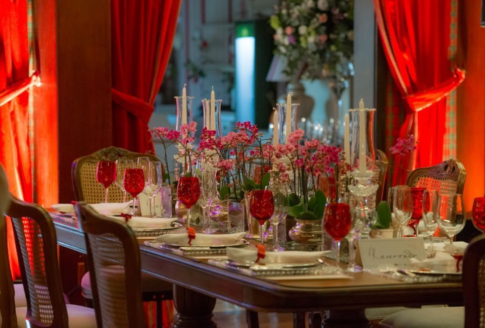 Mesa de jantar elegante com arranjos florais vermelhos, velas e cortinas vermelhas em uma sala formal.