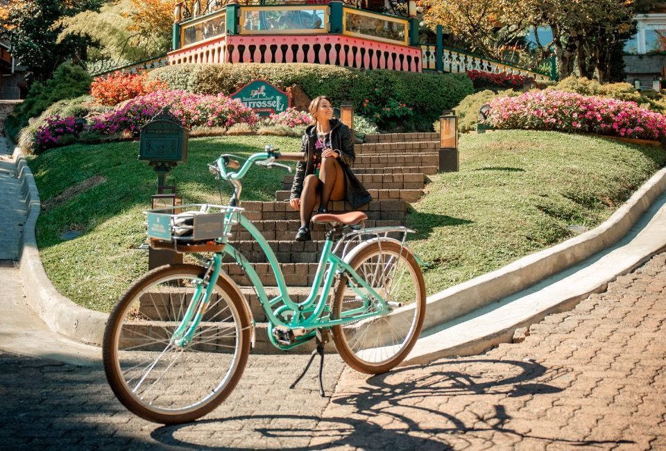 Bicicleta verde-menta estacionada em um parque ensolarado e florido, perto de degraus de pedra e uma ponte.