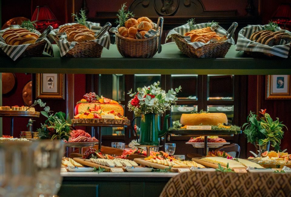 Buffet com cestas de pão, doces, frutas e flores sobre uma mesa de jantar decorada.