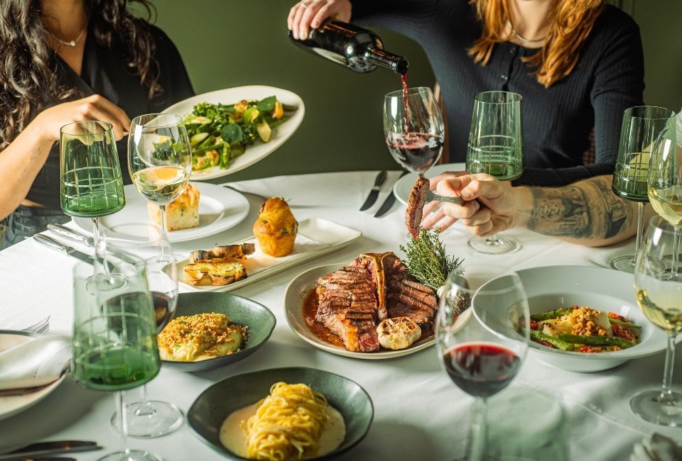 Pessoas compartilhando uma refeição em uma mesa de jantar com carne grelhada, massa, salada e taças de vinho.
