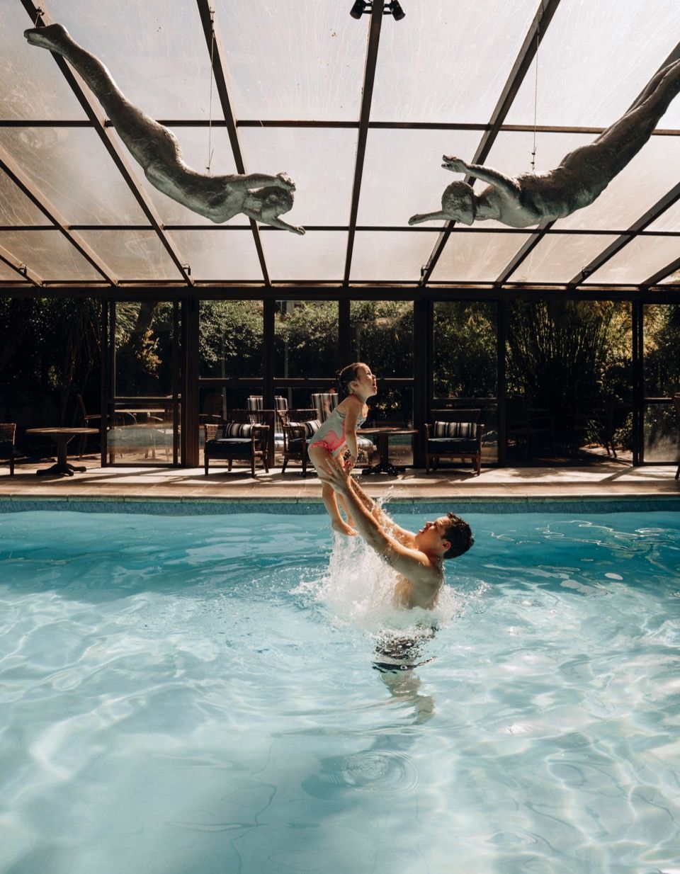 Cena de piscina coberta com uma pessoa levantando uma criança no ar, água espirrando sob um toldo iluminado pelo sol.
