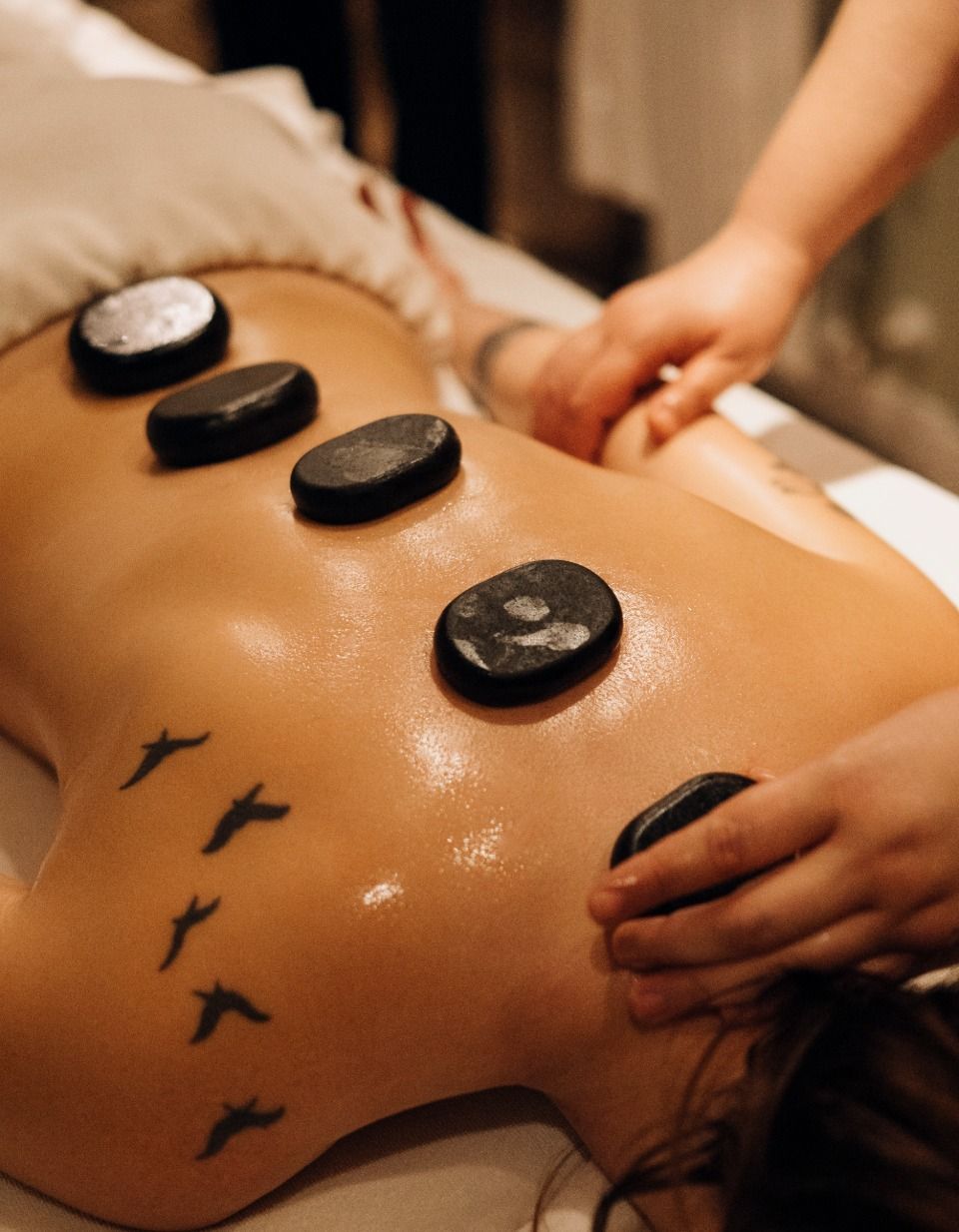Massagem com pedras quentes nas costas do cliente, utilizando pedras pretas lisas e óleo morno, com as mãos massageando a pele.