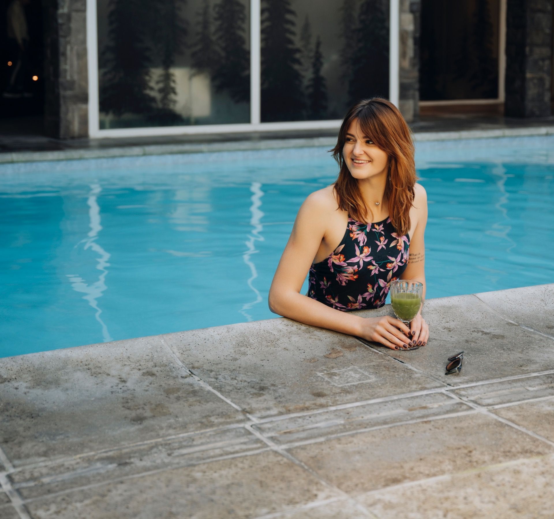 Mulher sentada à beira de uma piscina, segurando uma bebida, com uma expressão sorridente e árvores refletidas ao fundo.