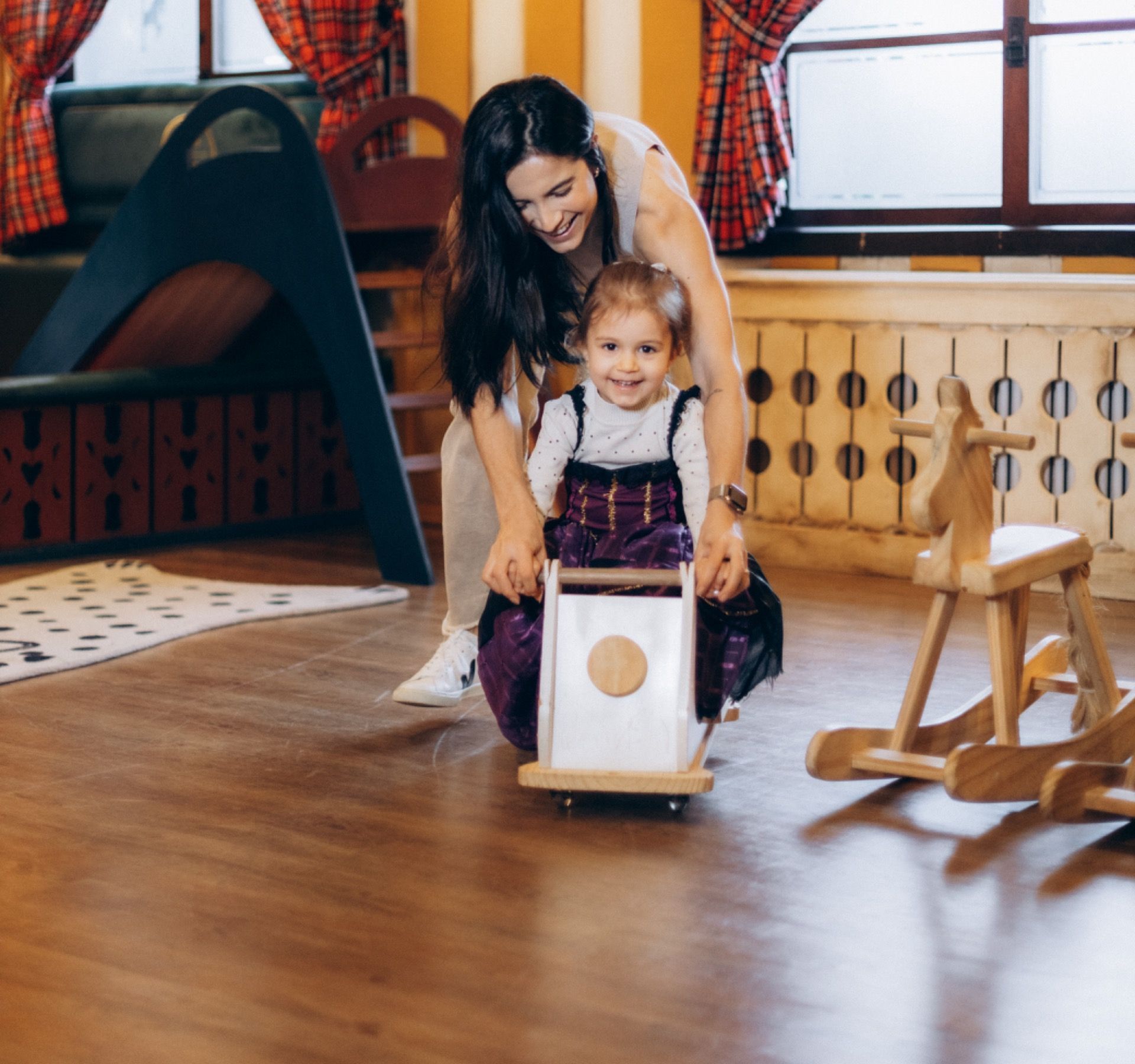 Mulher empurrando um andador de madeira com uma criança pequena em uma aconchegante sala de jogos interna, repleta de brinquedos e uma tenda indígena.