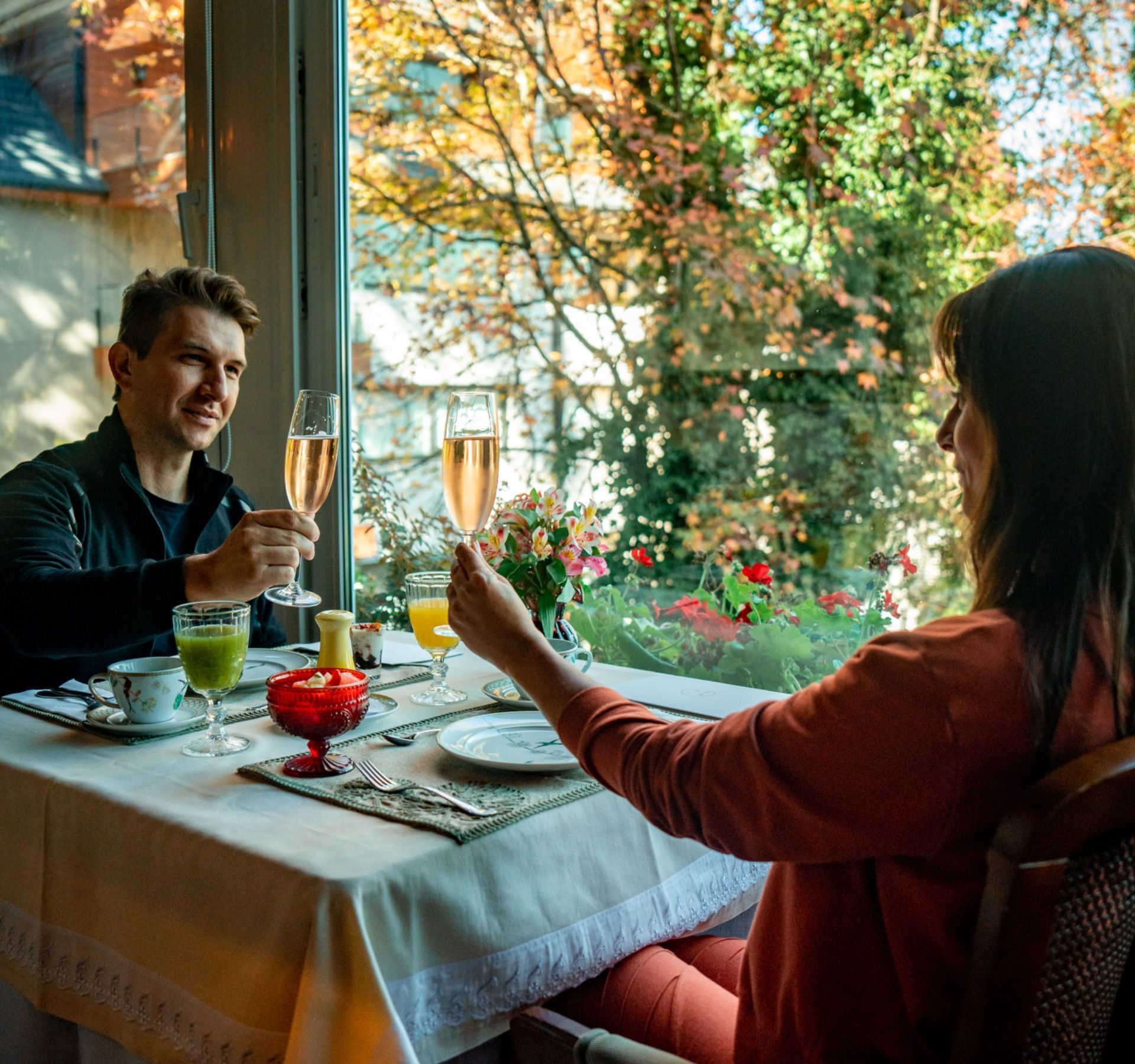 Duas pessoas brindando com champanhe em uma mesa aconchegante de restaurante perto de uma janela com vista para o jardim.