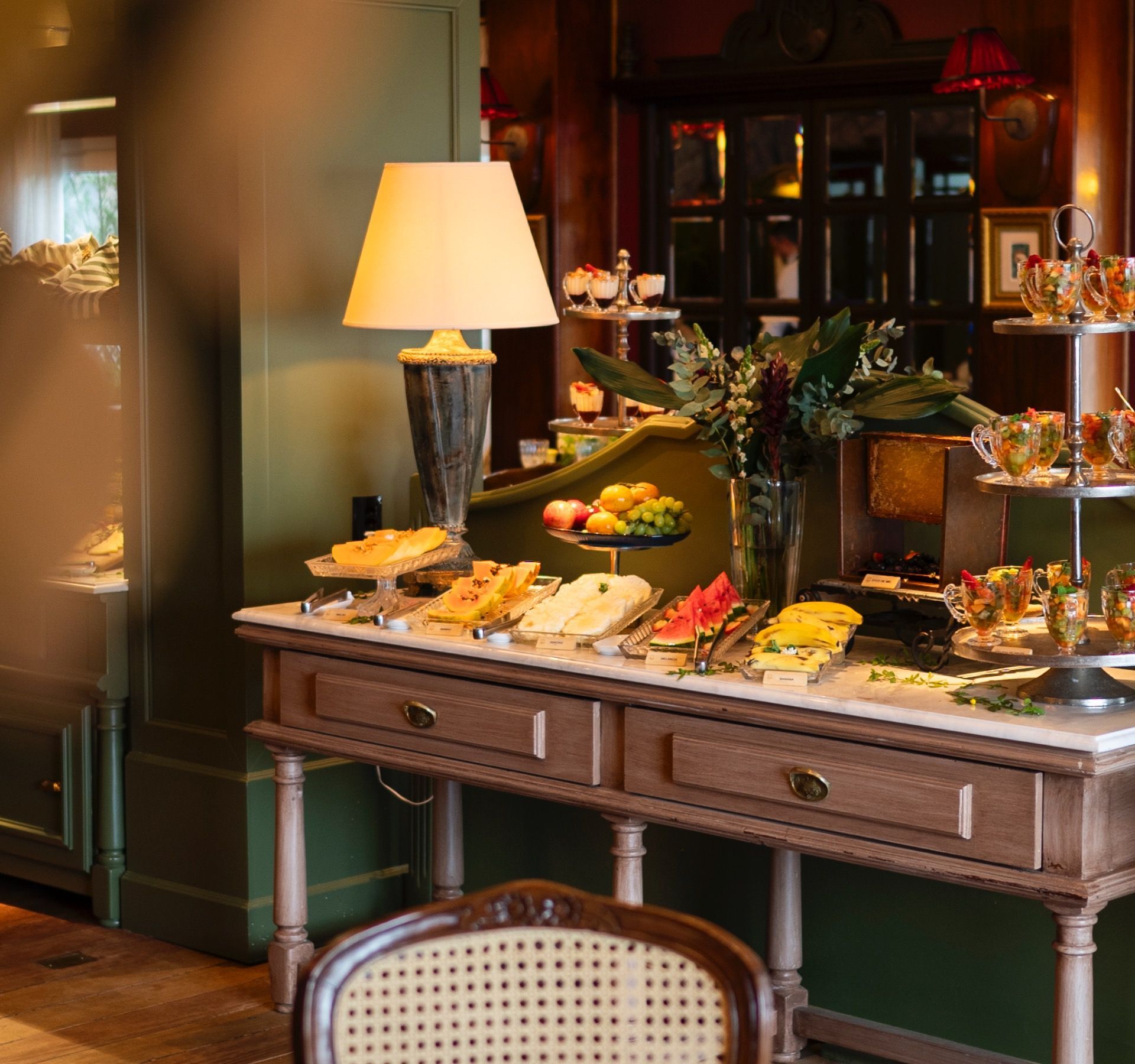 Elegante mesa de buffet com queijos, frutas e doces em uma sala de jantar com iluminação aconchegante.