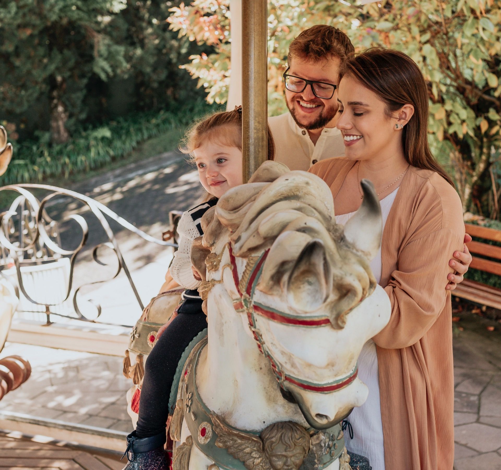 Família sorrindo em um cavalo de carrossel branco em um parque ensolarado, com um cachorro em primeiro plano.