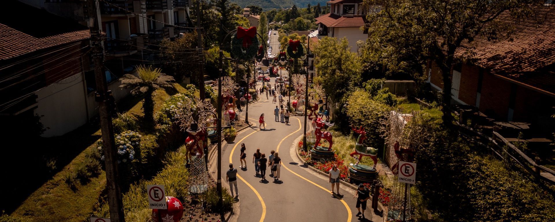 Rua de pedestres movimentada, com pessoas caminhando por um caminho sinuoso em uma área ensolarada e arborizada da cidade.